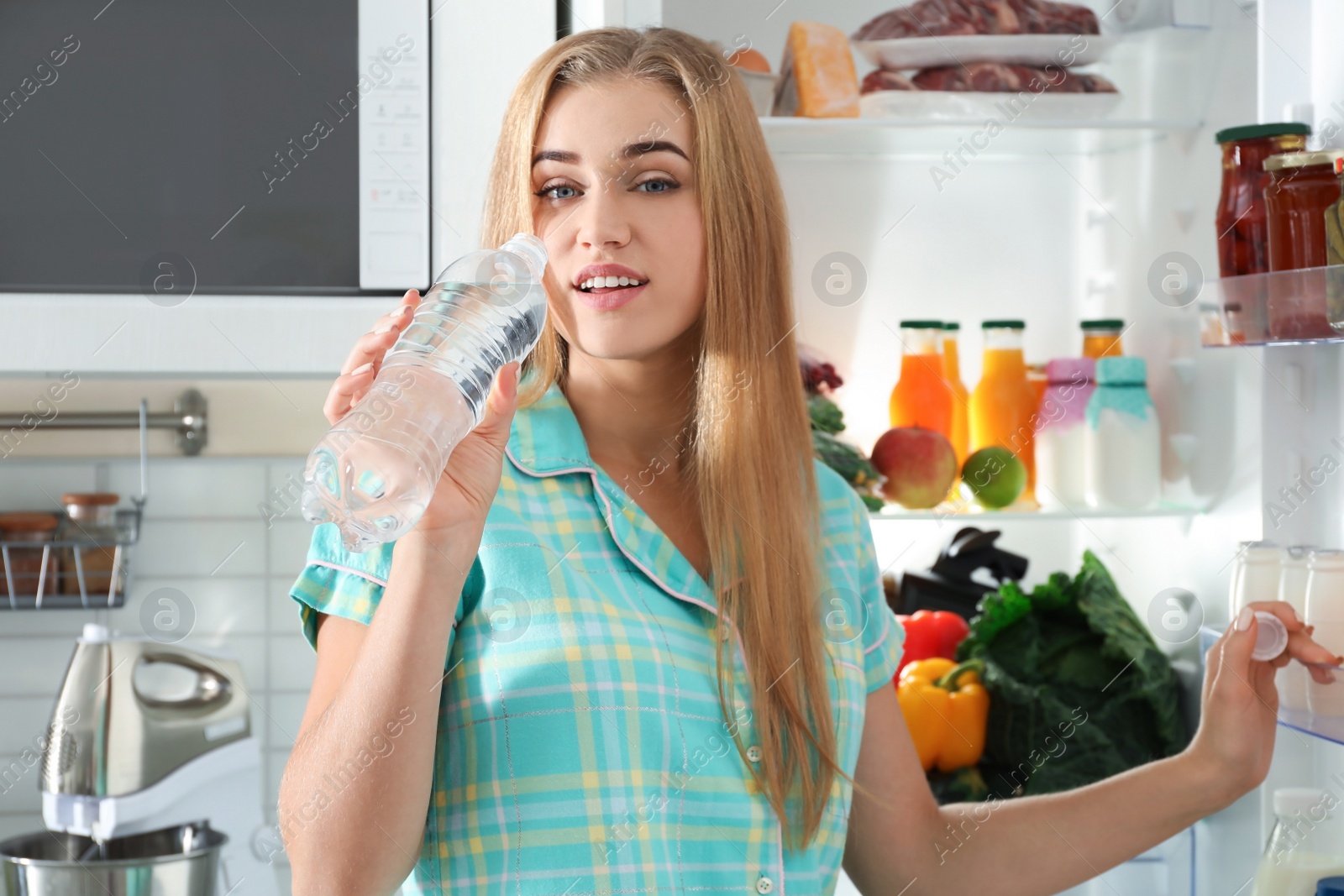 Photo of Woman drinking water out of bottle near refrigerator in kitchen