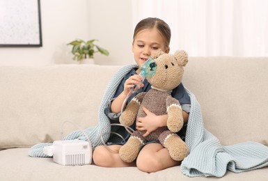 Little girl with toy bear and nebulizer for inhalation on sofa at home