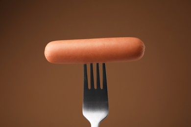 Delicious boiled sausage on fork against brown background, closeup