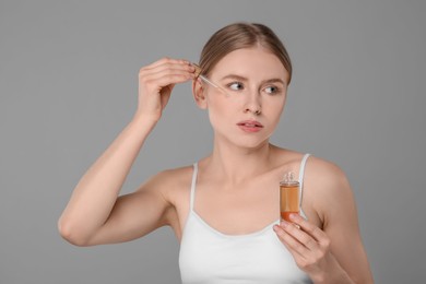 Beautiful young woman applying essential oil onto face on grey background