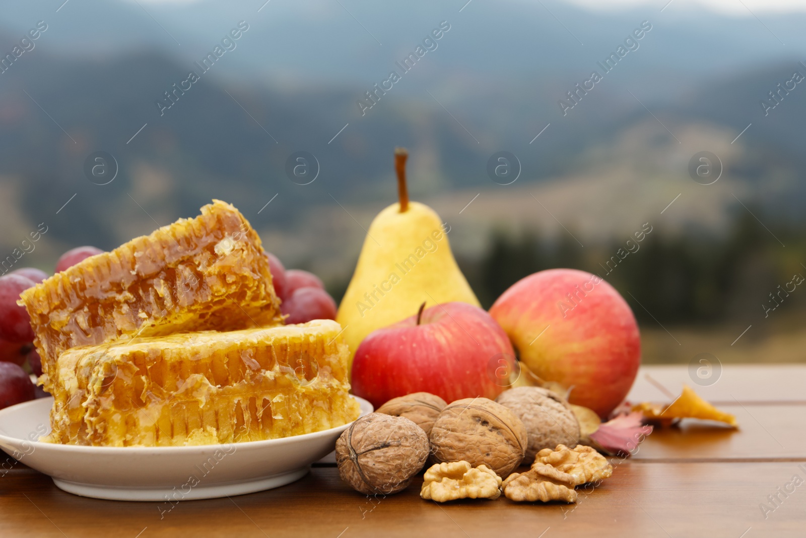 Photo of Tasty fresh honeycombs, fruits and walnuts on wooden table against mountain landscape