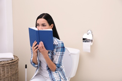 Woman with book sitting on toilet bowl in bathroom