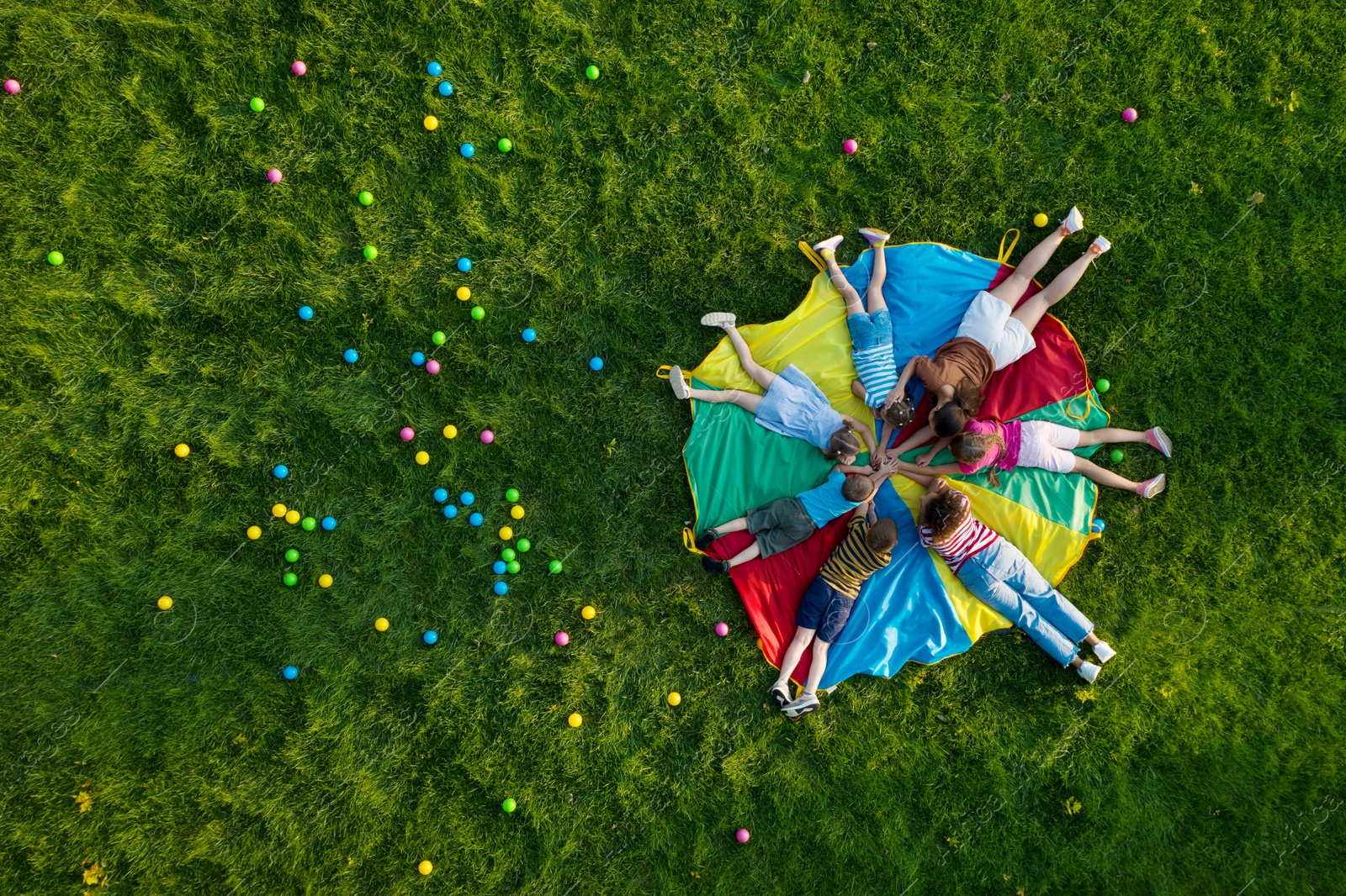 Image of Group of children with teachers holding hands together on rainbow playground parachute in park, top view. Summer camp activity