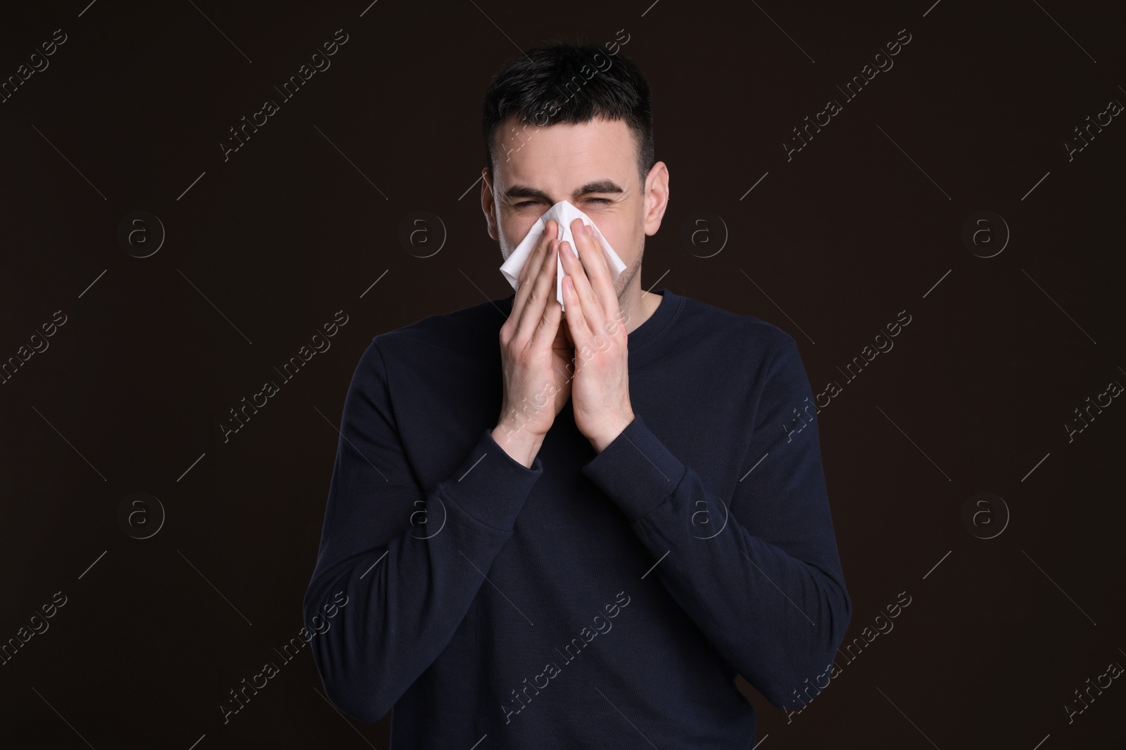 Photo of Young man blowing nose in tissue on dark background. Cold symptoms