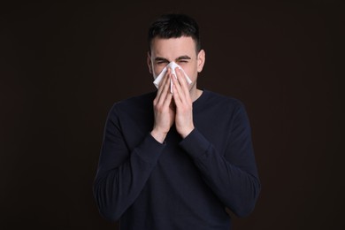 Photo of Young man blowing nose in tissue on dark background. Cold symptoms