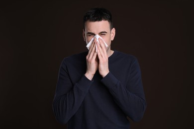 Young man blowing nose in tissue on dark background. Cold symptoms