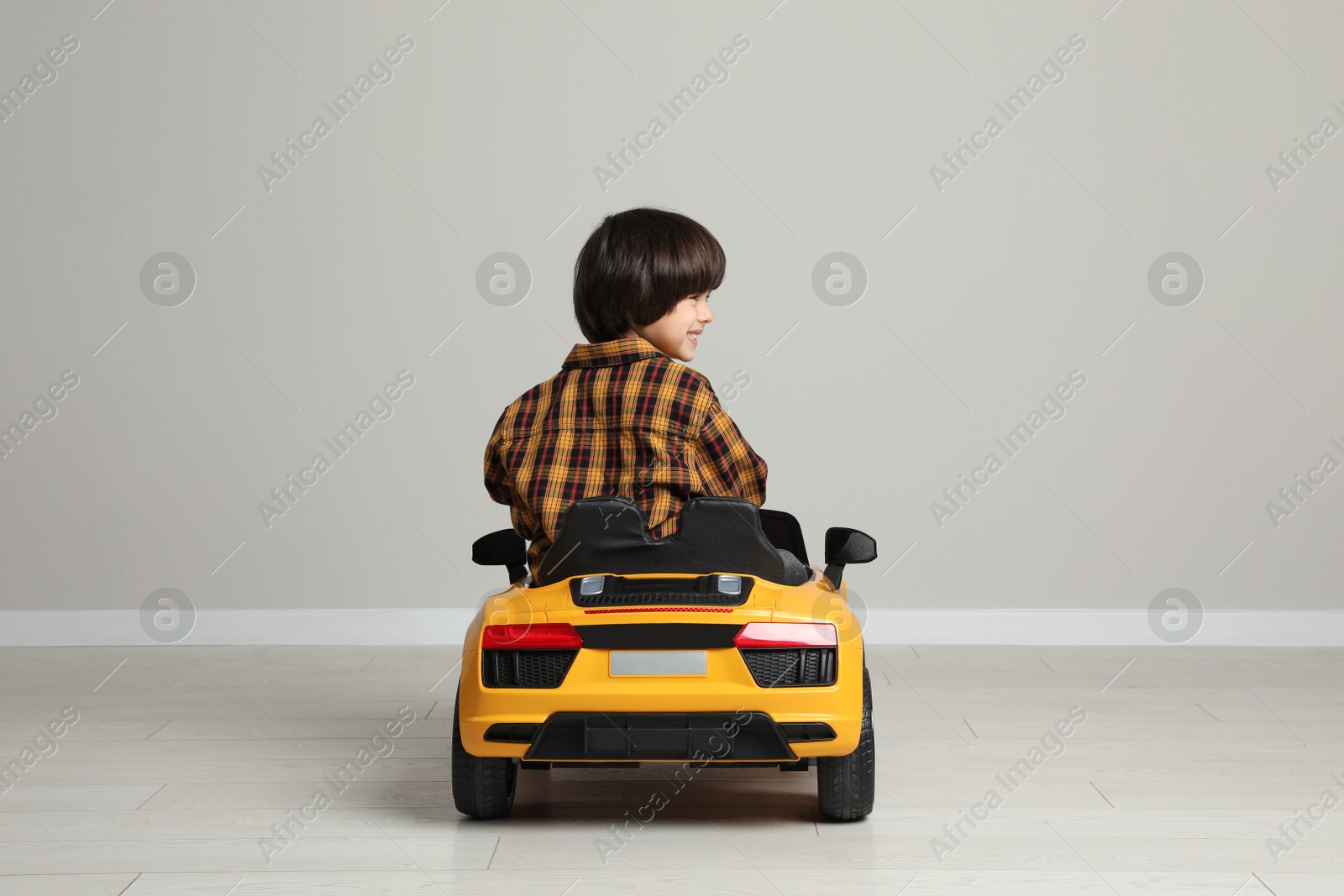 Photo of Cute little boy driving children's electric toy car near grey wall indoors, back view