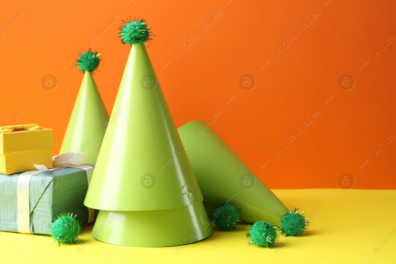 Photo of Party hats and gift boxes on yellow table against orange background, space for text