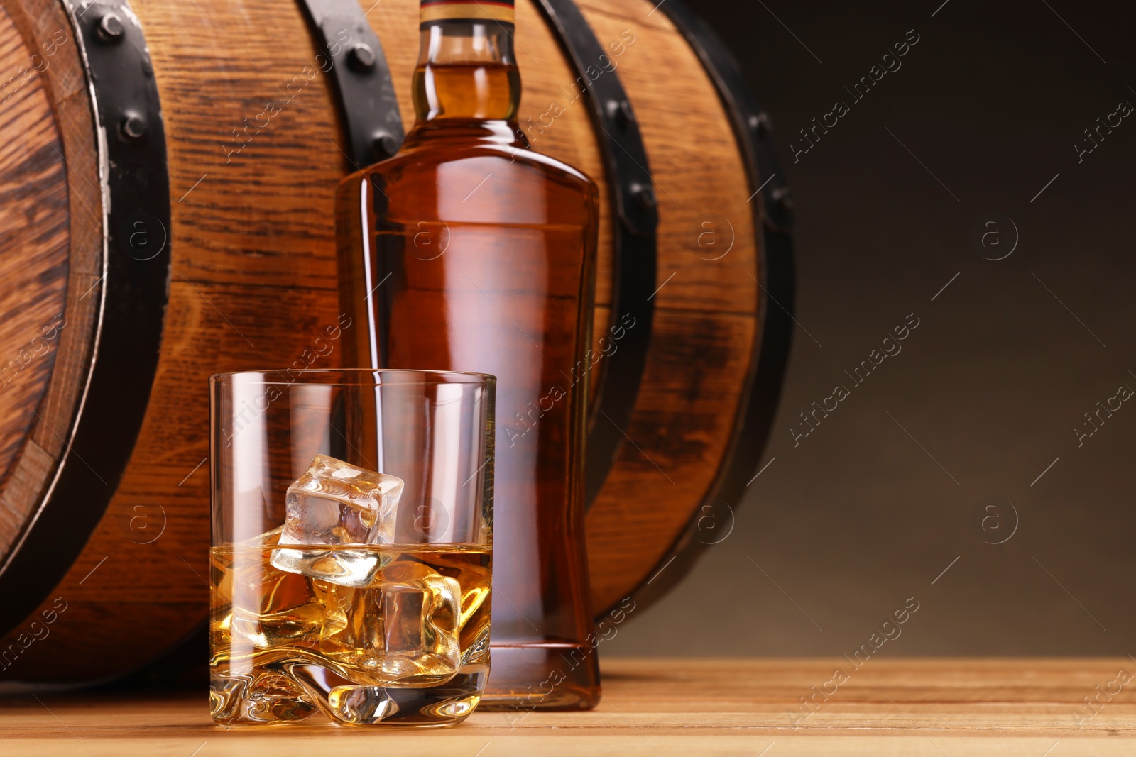 Photo of Whiskey with ice cubes in glass, bottle and barrel on wooden table against dark background, space for text