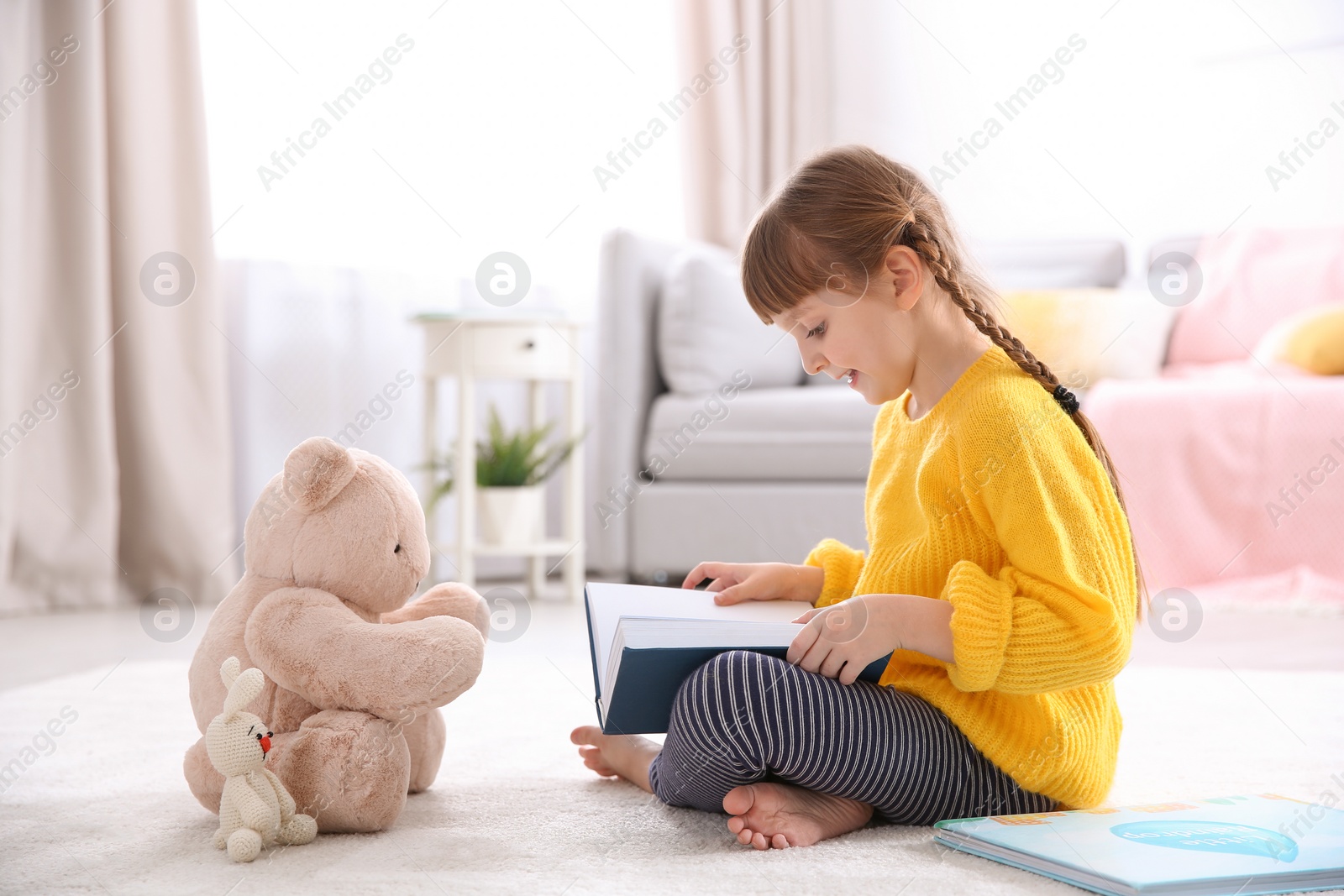 Photo of Cute little girl with teddy bear reading book on floor at home