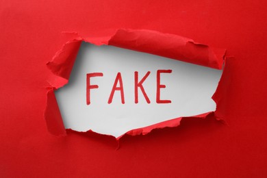 Photo of Word Fake written on white background, view through hole in red torn paper