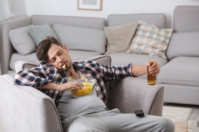 Photo of Lazy man with bottle of beer and chips sleeping in armchair at home