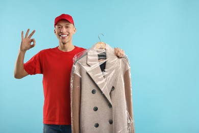 Photo of Dry-cleaning delivery. Happy courier holding coat in plastic bag and showing OK gesture on light blue background, space for text