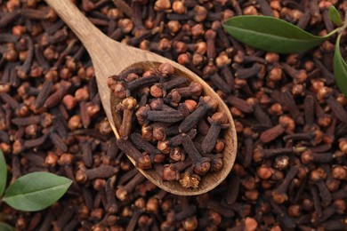 Many aromatic cloves, wooden spoon and green leaves as background, top view