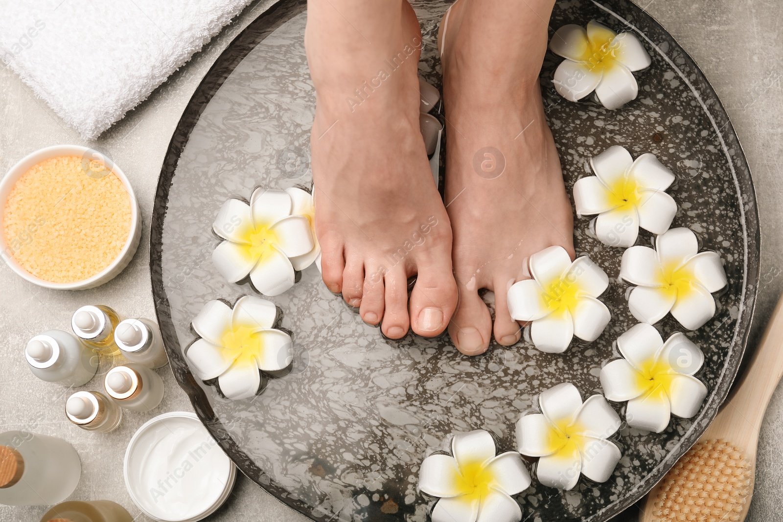 Photo of Woman soaking her feet in bowl with water and flowers on light grey floor, top view. Spa treatment