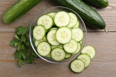 Photo of Cut cucumber in glass bowl, fresh vegetables and parsley on wooden table, flat lay