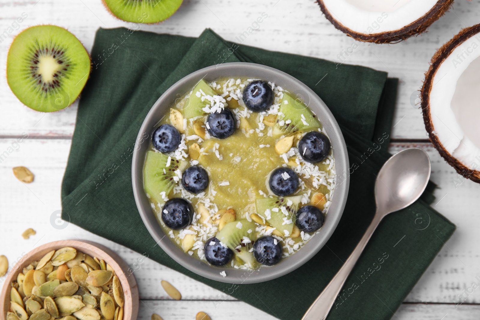 Photo of Bowl of delicious fruit smoothie served with fresh blueberries, kiwi slices and coconut flakes on white wooden table, flat lay