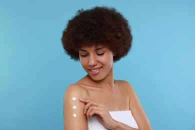 Beautiful young woman applying body cream onto arm on light blue background