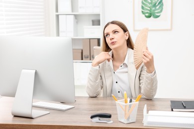 Photo of Businesswoman waving hand fan to cool herself at table in office