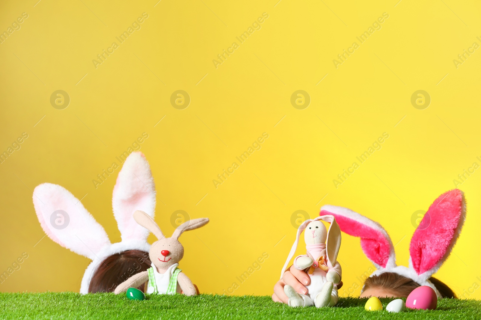 Photo of Little girls wearing rabbit ears headbands and playing with toy Easter bunnies on green grass surface against color background