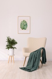 Comfortable armchair, blanket, houseplant and picture on white wall indoors