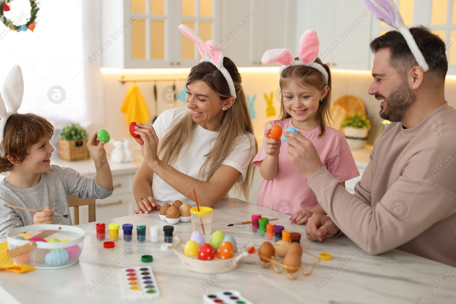 Photo of Easter celebration. Happy family with bunny ears having fun while painting eggs at white marble table in kitchen