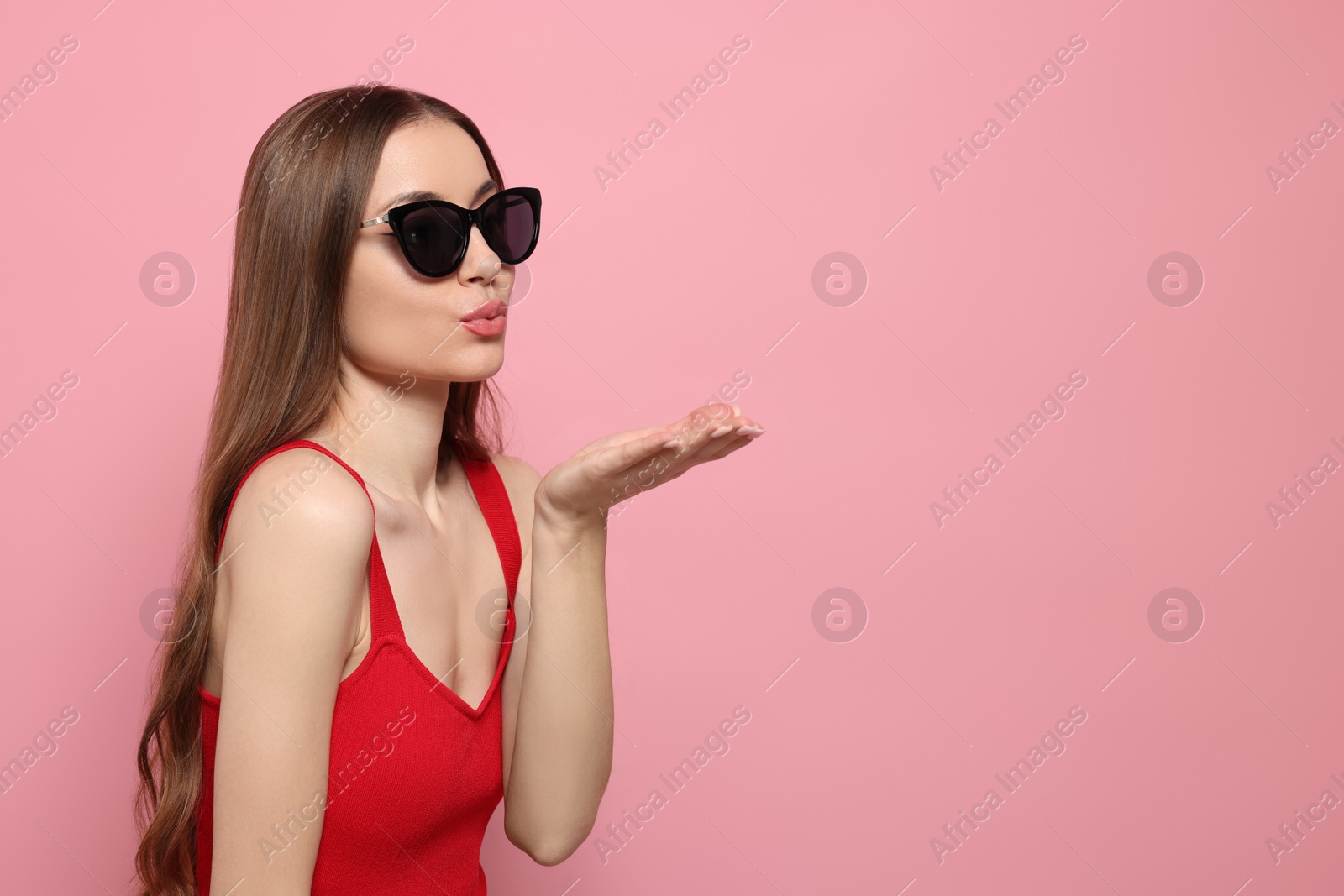 Photo of Beautiful young woman with sunglasses blowing kiss on pink background, space for text