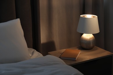 Stylish lamp and book on bedside table near bed indoors