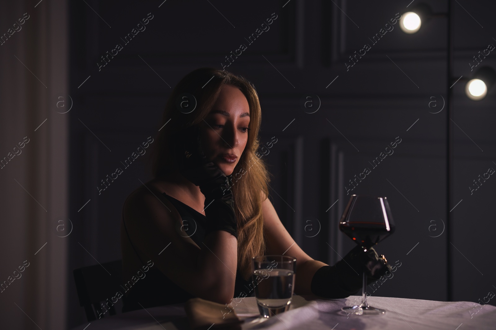 Photo of Elegant young woman with glass of wine at table indoors in evening