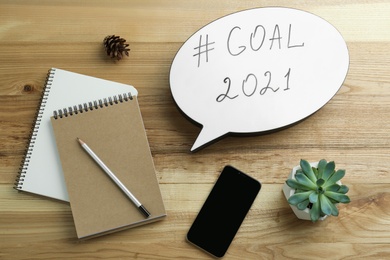 Photo of Paper speech bubble with hashtag Goal 2021 near notebooks and smartphone on wooden table, flat lay. New year targets