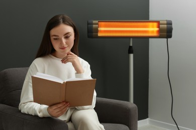 Photo of Young woman reading book on armchair near electric infrared heater at home. Space for text