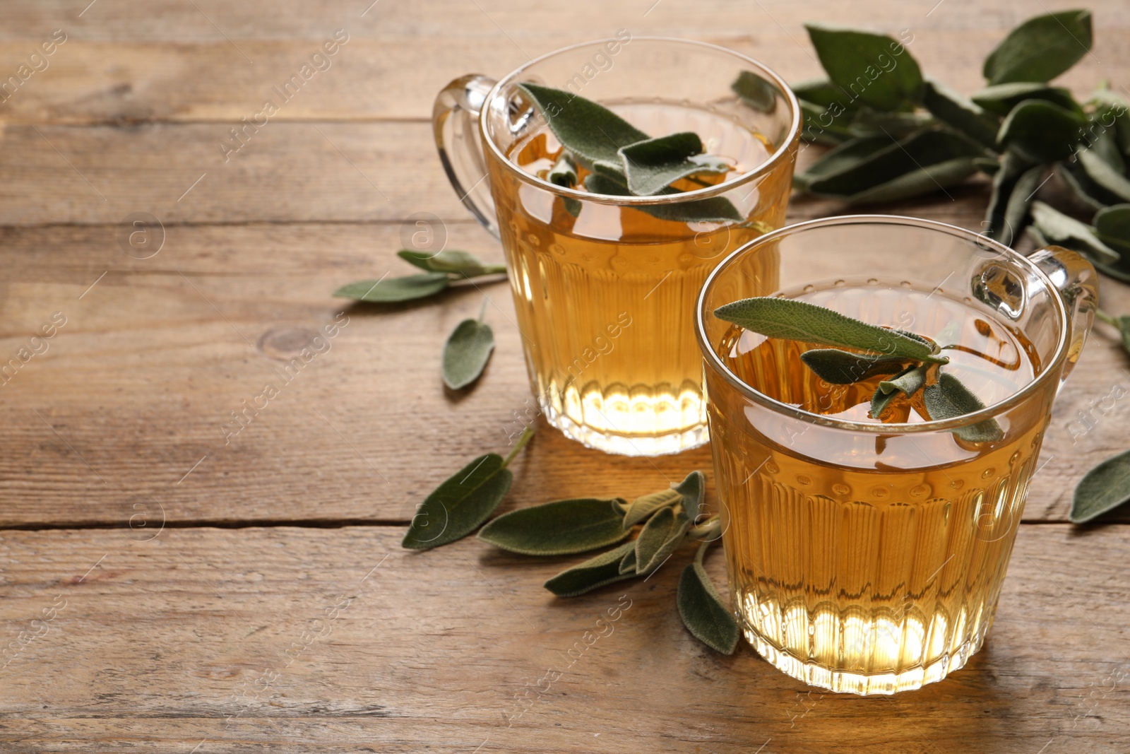 Photo of Cups of aromatic sage tea and fresh leaves on wooden table. Space for text