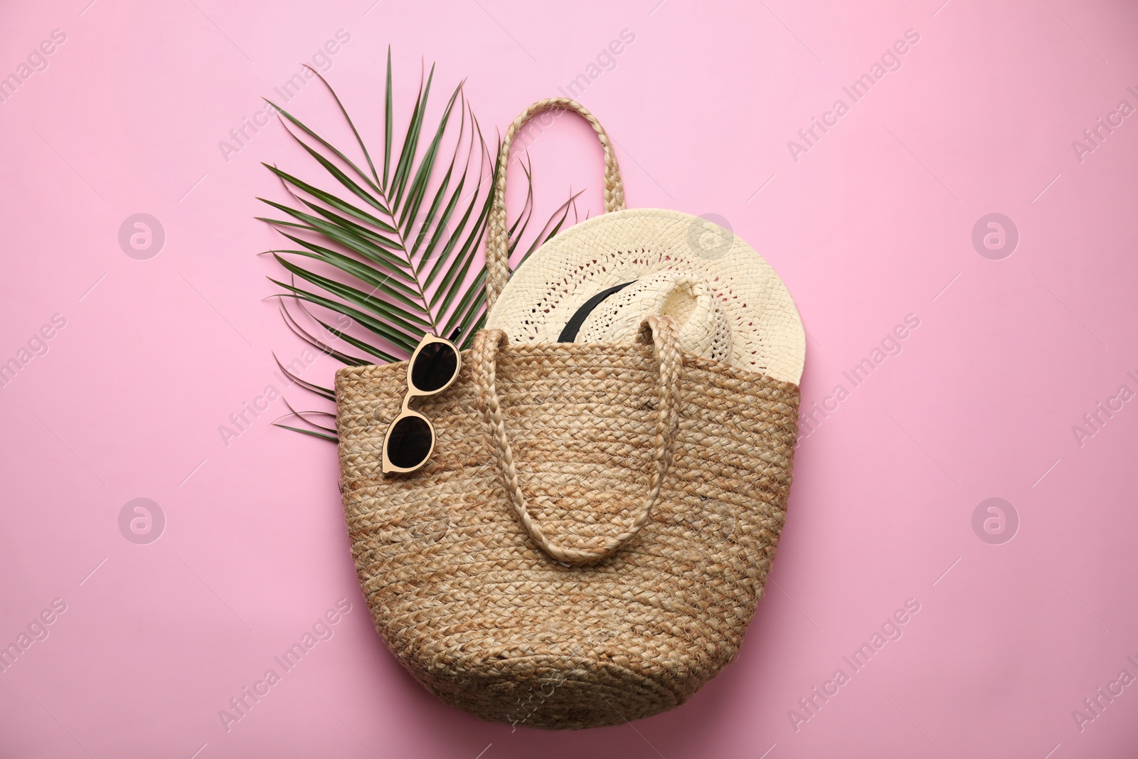 Photo of Elegant woman's straw bag with hat, tropical leaf and sunglasses on pink background, top view