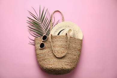 Elegant woman's straw bag with hat, tropical leaf and sunglasses on pink background, top view