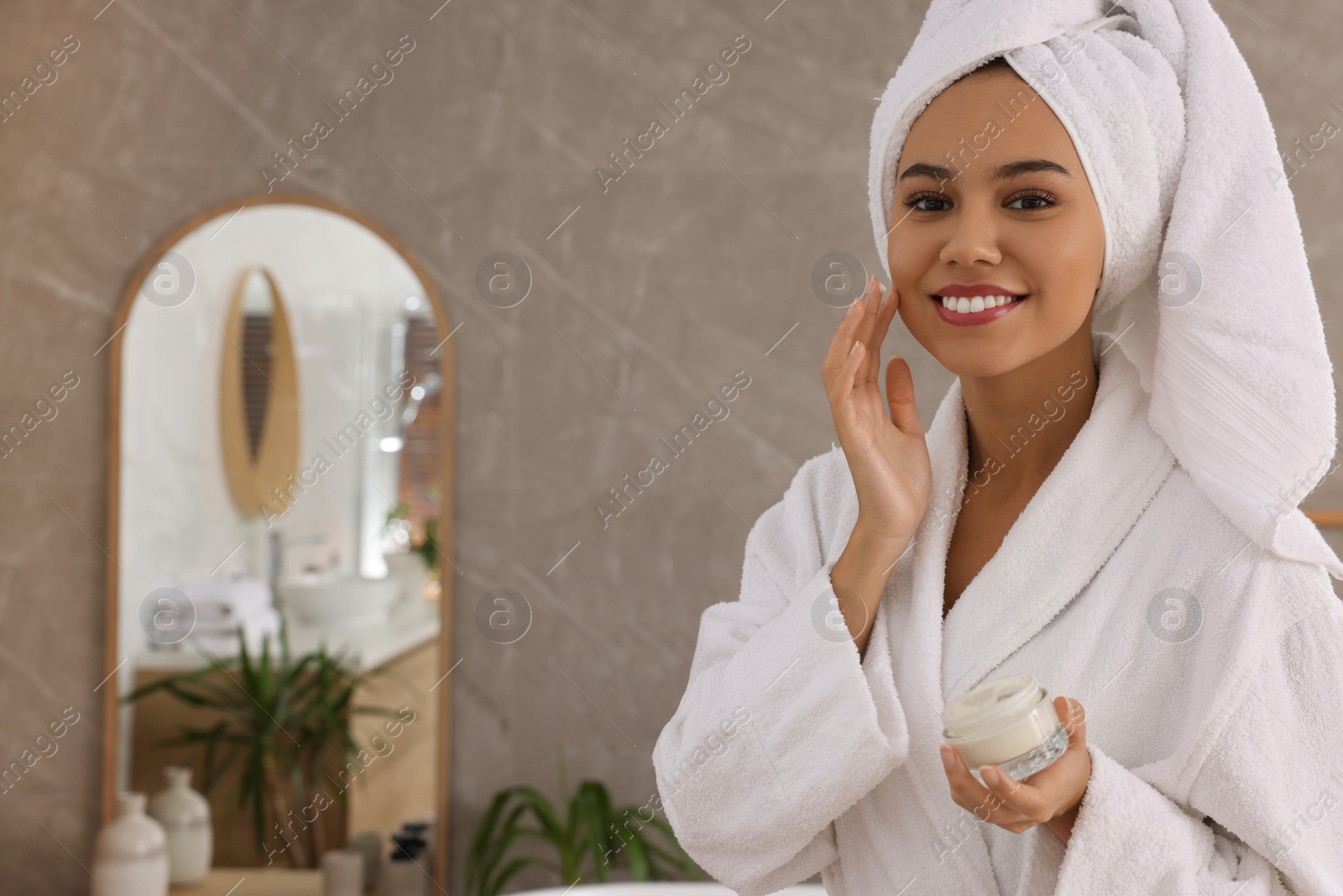 Photo of Beautiful African American woman applying cream onto face in bathroom, space for text