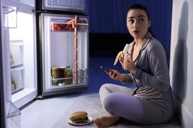 Young woman eating banana while using smartphone near fridge in kitchen at night. Bad habit