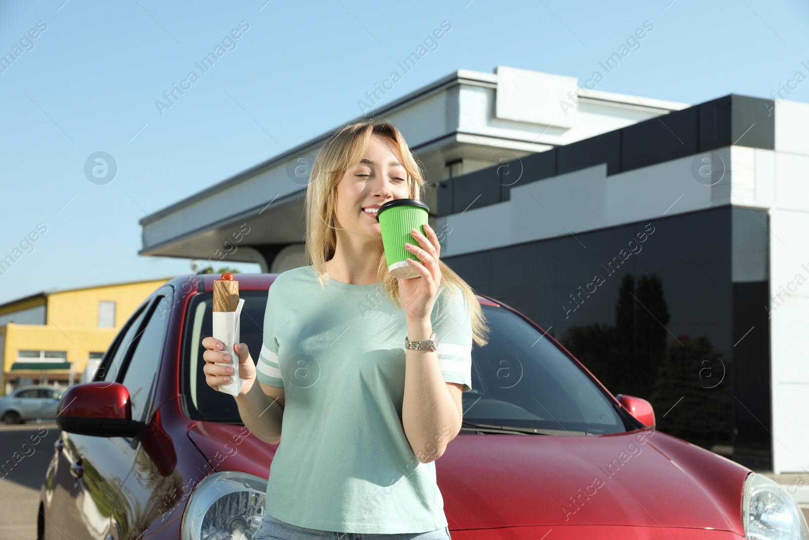 Photo of Beautiful young woman with hot dog drinking coffee near car at gas station