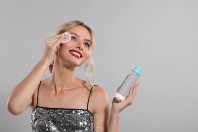 Smiling woman removing makeup with cotton pad and holding bottle on light grey background. Space for text
