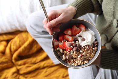 Woman eating tasty granola with chocolate chips, strawberries and yogurt indoors, top view