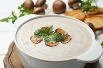 Fresh homemade mushroom soup in ceramic pot on white wooden table, closeup