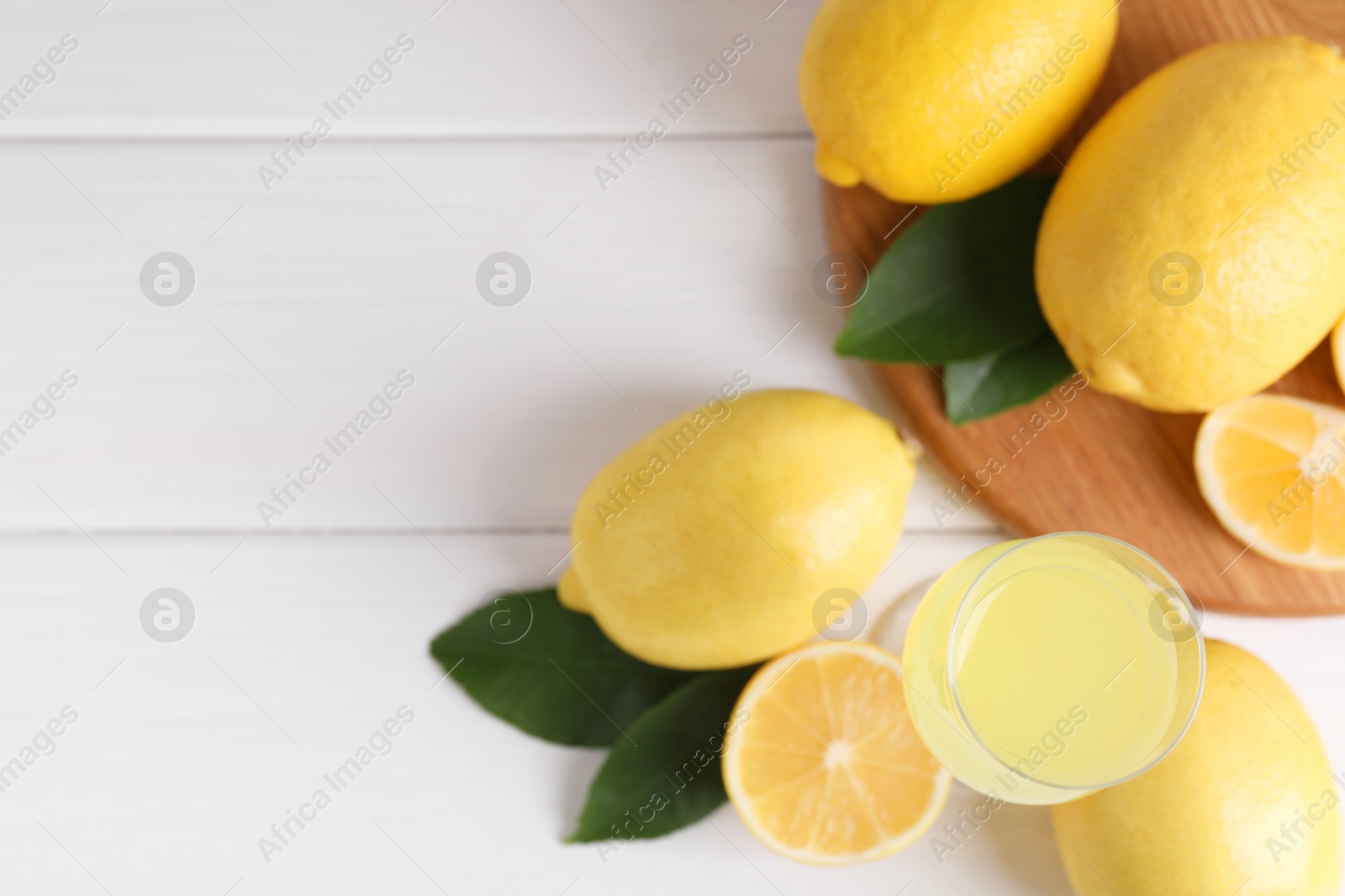 Photo of Liqueur glass with tasty limoncello, lemons and green leaves on white wooden table, flat lay. Space for text