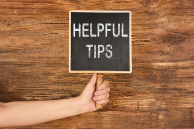 Image of Woman holding small chalkboard with text Helpful Tips on wooden background, closeup