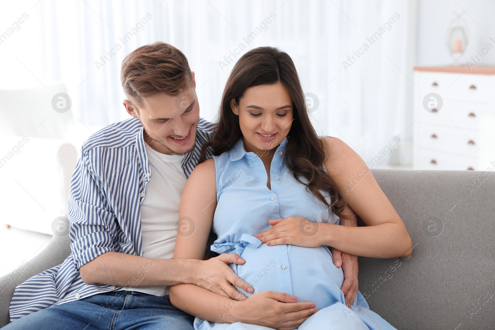 Photo of Young man and his pregnant wife sitting on sofa at home