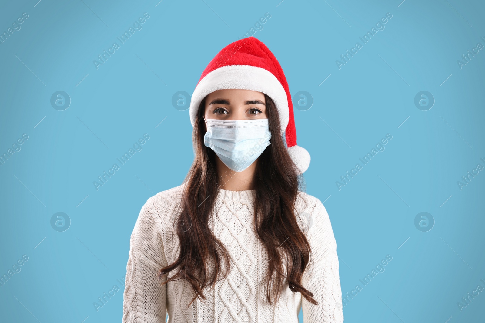 Photo of Beautiful woman wearing Santa Claus hat and medical mask on light blue background