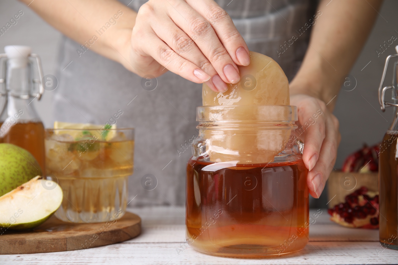 Photo of Woman putting Scoby fungus into jar with kombucha at white wooden table, closeup