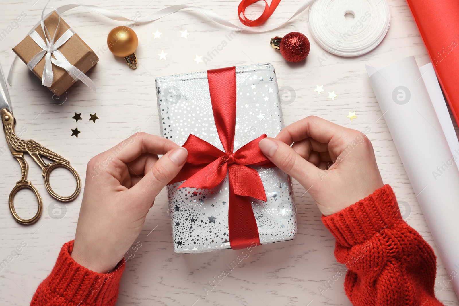 Photo of Christmas present. Woman tying ribbon bow on gift box at white wooden table, top view