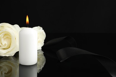 Photo of Burning candle, white roses and ribbon on black mirror surface in darkness, closeup with space for text. Funeral symbols