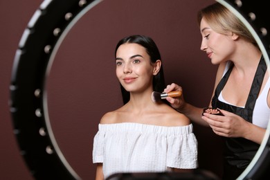 Photo of Professional makeup artist working with beautiful young woman against brown background, view through ring lamp