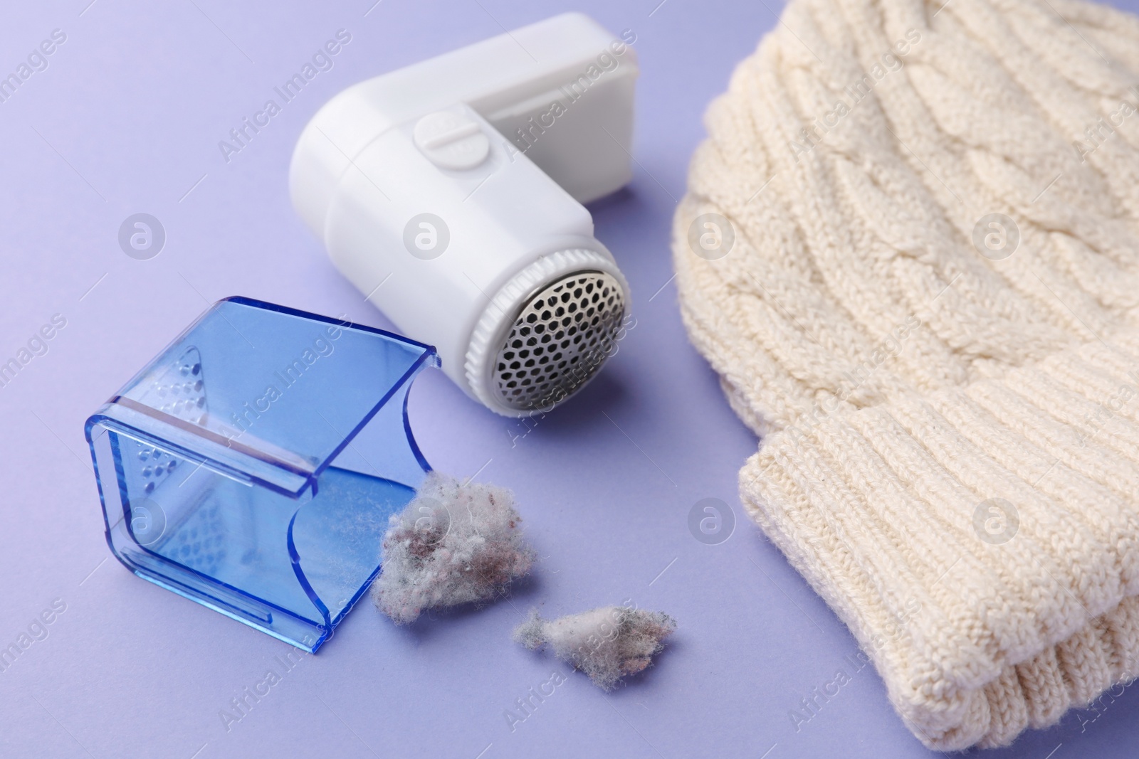 Photo of Fabric shaver with fuzz and knitted hat on lilac background, closeup