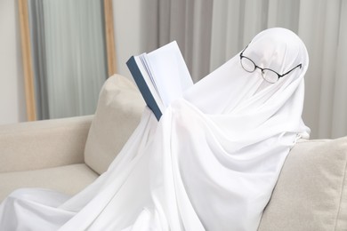 Photo of Creepy ghost. Person covered with white sheet reading book on sofa at home